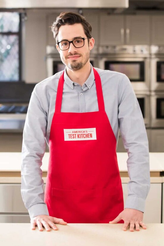 man standing at a counter in a red apron that reads "America's Test Kitchen."