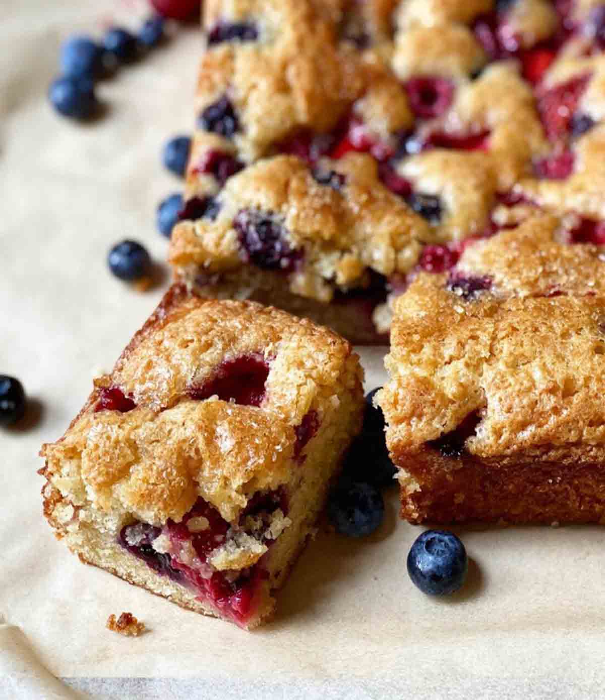 A rectangular easy mixed berry snacking cake with one piece cut from it and blueberries on the side.