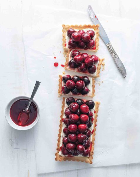 A rectangular fresh cherry tart with cream cheese filling on a white surface with a knife and a small bowl of jam on the side.