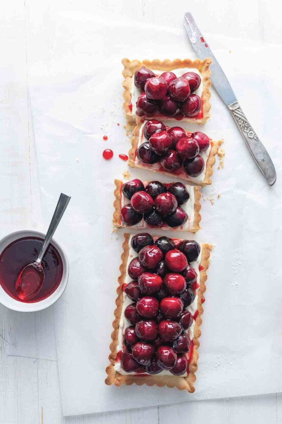A rectangular fresh cherry tart with cream cheese filling on a white surface with a knife and a small bowl of jam on the side.