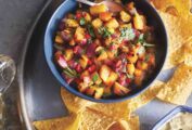 A blue bowl filled with grilled pineapple salsa on a platter with tortilla chips., beer, and lime wedges.