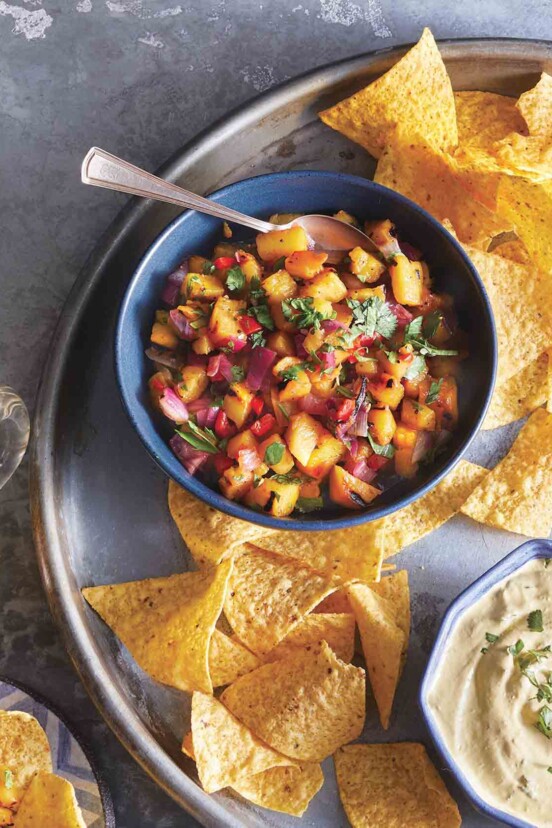 A blue bowl filled with grilled pineapple salsa on a platter with tortilla chips., beer, and lime wedges.