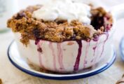 A individual huckleberry crisp in a white bowl topped with whipped cream and a spoon on the side.