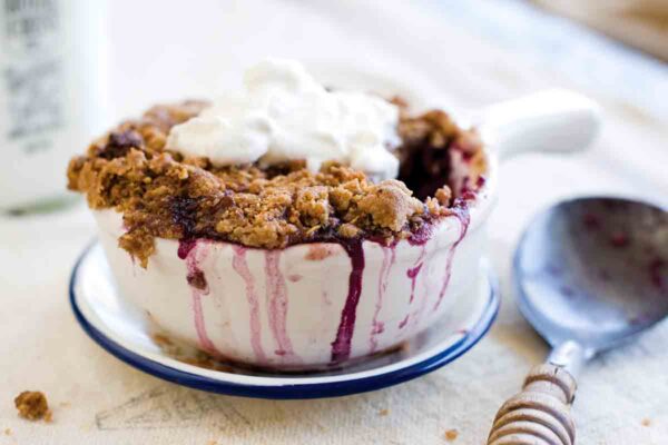 A individual huckleberry crisp in a white bowl topped with whipped cream and a spoon on the side.
