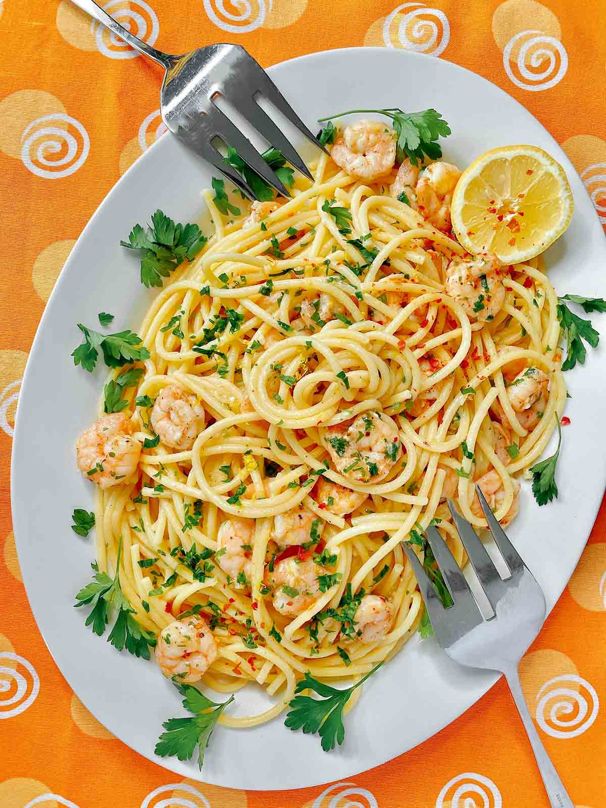 A white platter filled with lemon garlic shrimp pasta, garnished with parsley and a lemon wedge and two serving forks resting on the platter.