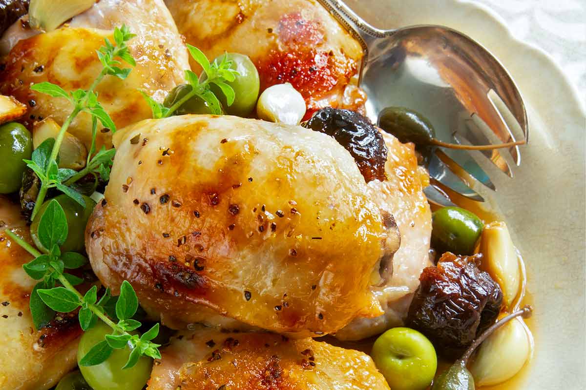 Pieces of chicken Marbella in a white serving dish with prunes, olives, and capers, garnished with fresh thyme and a serving spoon on the side.