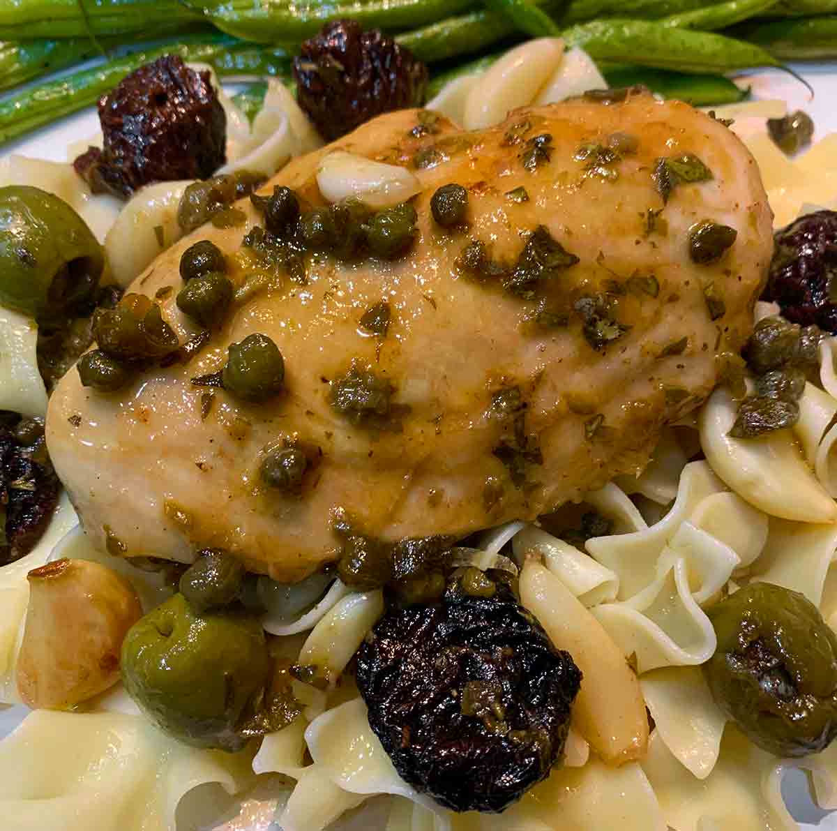 A pieces of modern chicken Marbella on egg noodles with green beans on the side.