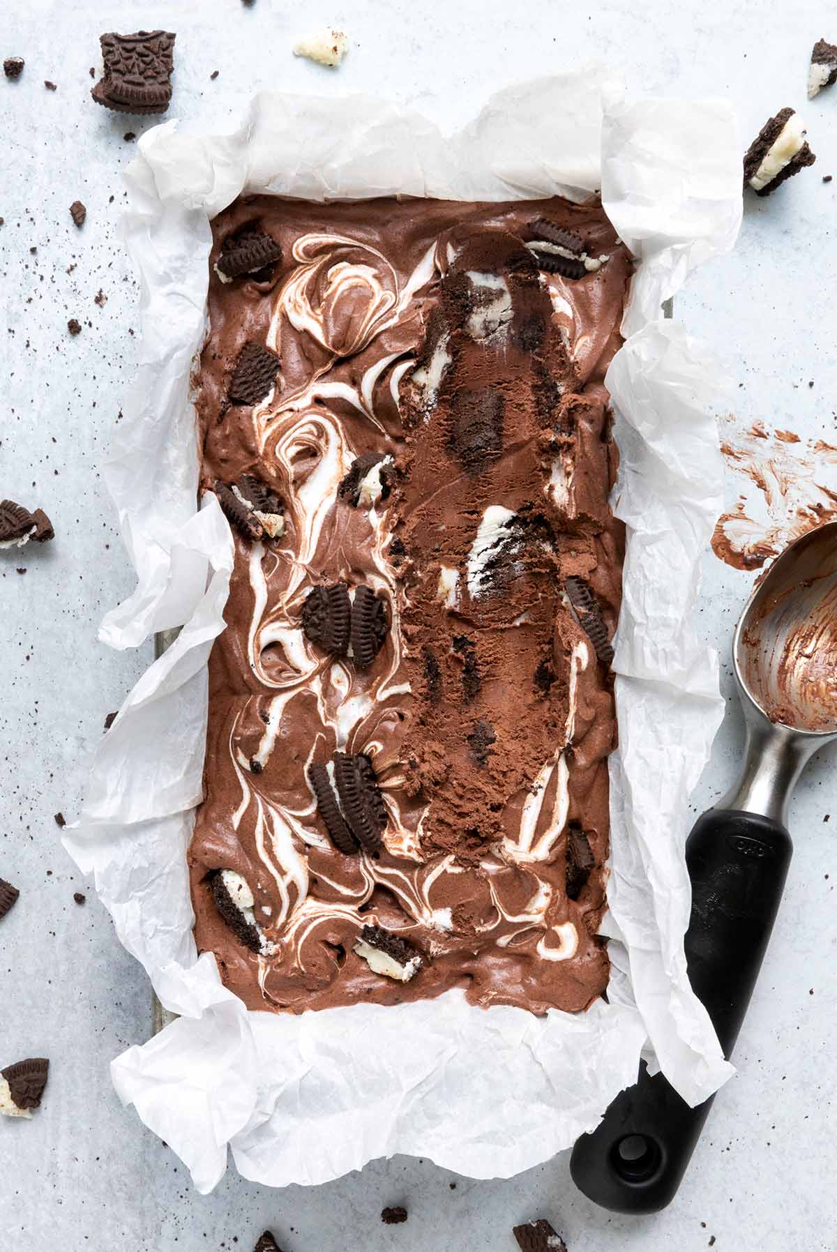A loaf pan filled with chocolate Oreo ice cream with pieces of Oreo scattered around it and a ice cream scoop on the side.