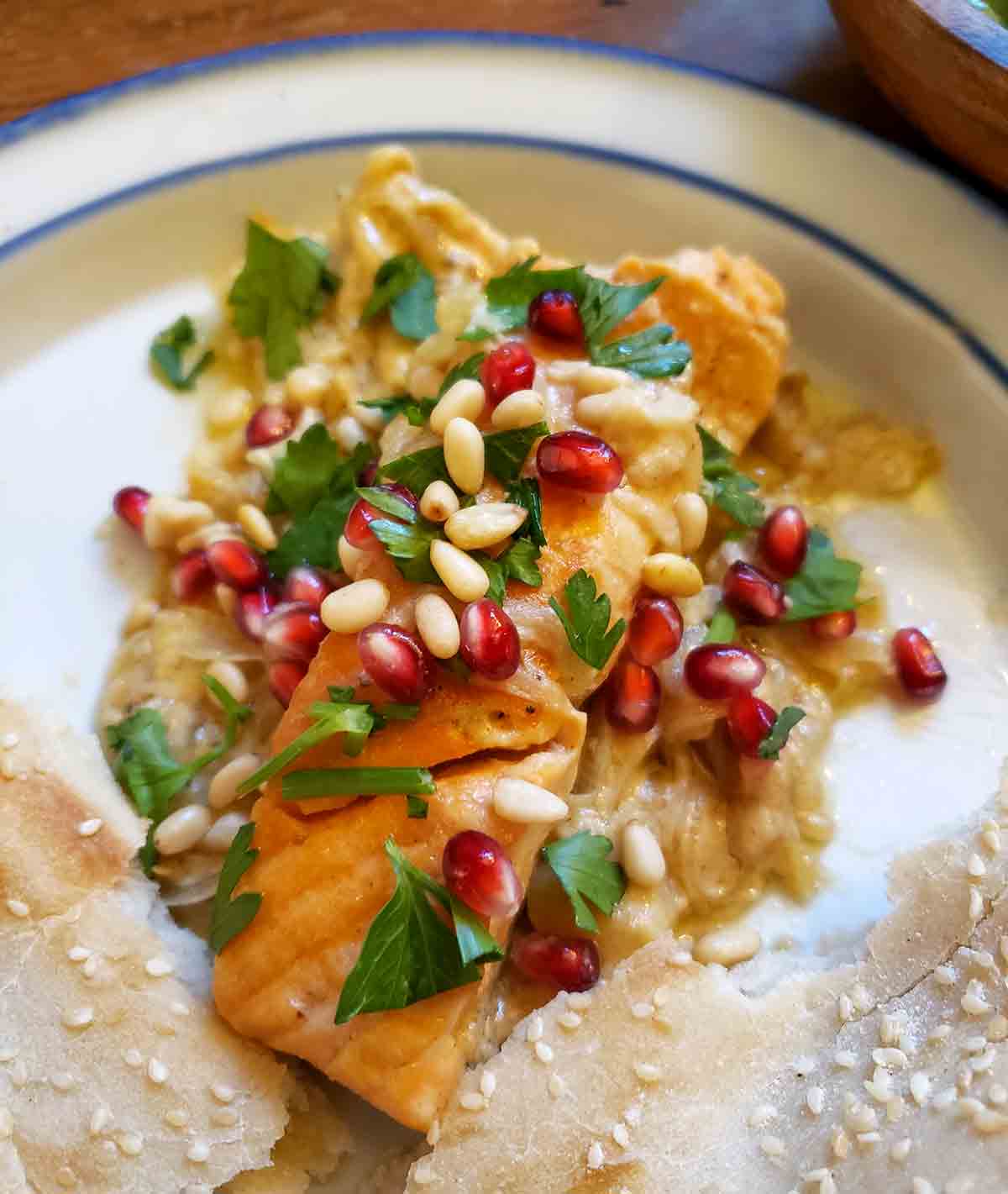 A piece of pan seared salmon with sweet tahini sauce, garnished with pine nuts, pomegranate seeds, and parsley on a white plate.