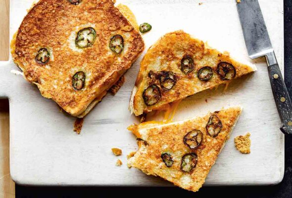 Two Parmesan-crusted grilled cheese with jalapeños on a white cutting board, one cut in half, with a knife lying beside them.