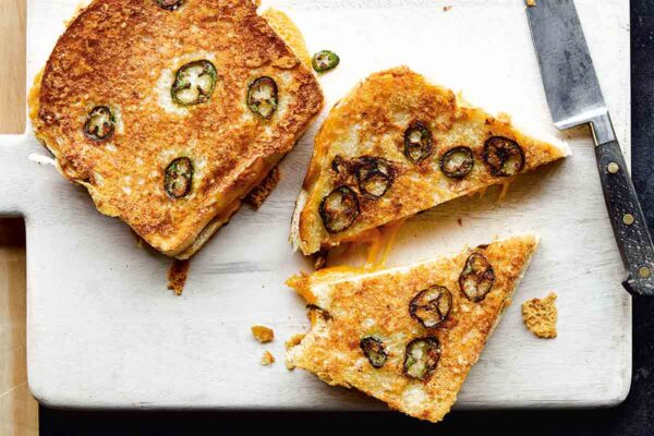 Two Parmesan-crusted grilled cheese with jalapeños on a white cutting board, one cut in half, with a knife lying beside them.