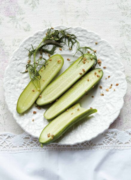 Three and a half quick dill pickles, dill seed, and a sprig of dill on a decorative white plate.
