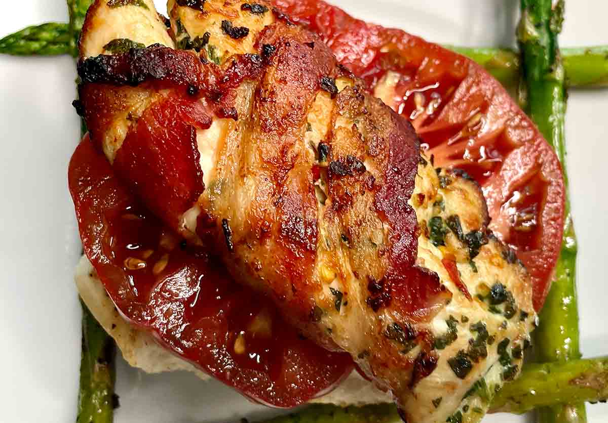 A spicy bacon-wrapped chicken breast on tomato slices on top of four asparagus spears.