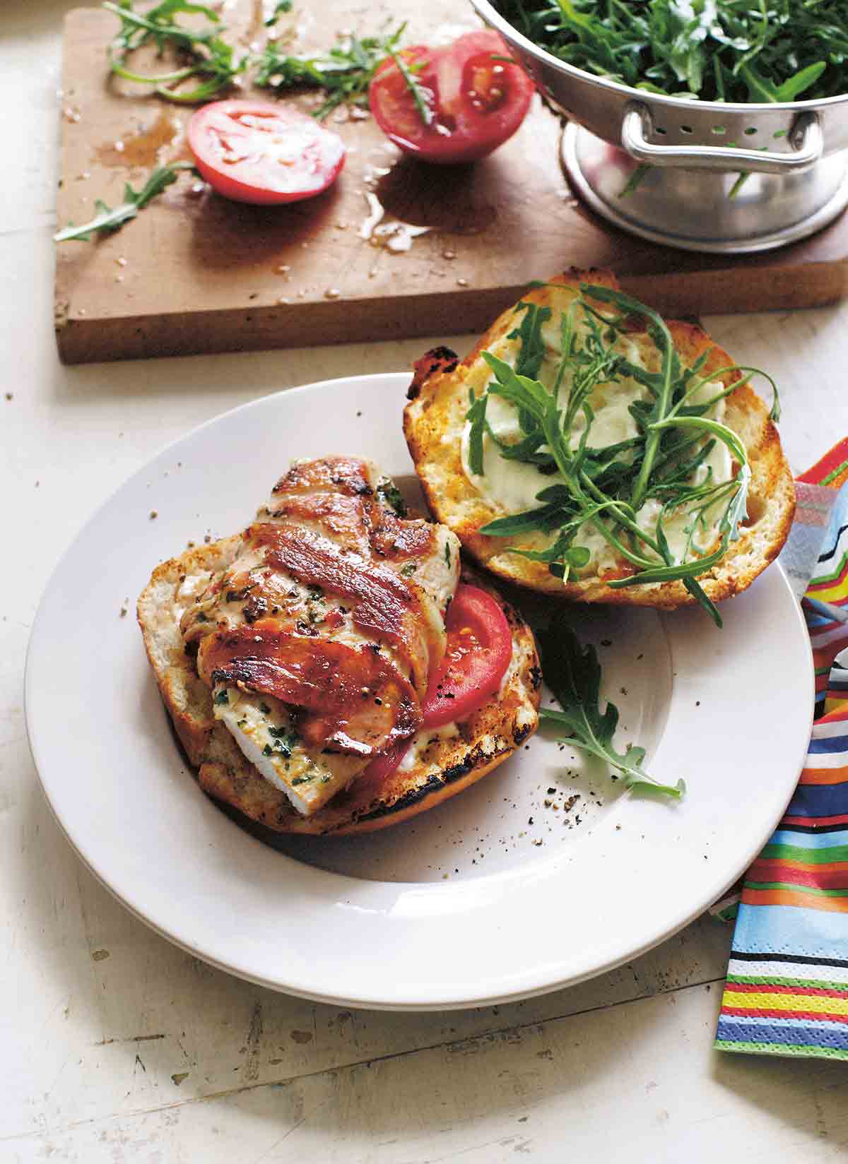 A spicy bacon-wrapped grilled chicken breast on top of a bun and tomato slices with the top half of the bun and some arugula next to it.