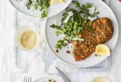 Three plates topped with pistachio-crusted chicken cutlets, arugula salad, and lemon wedges, with forks and knives on the side.