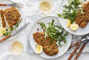 Three plates topped with pistachio-crusted chicken cutlets, arugula salad, and lemon wedges, with forks and knives on the side.