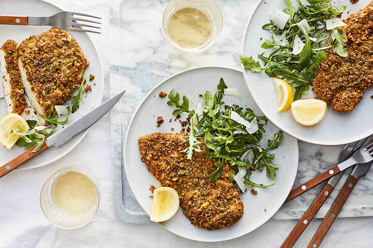 Three plates of pistachio-crusted chicken cutlets, arugula salad, and lemon wedges, served with forks and knives.