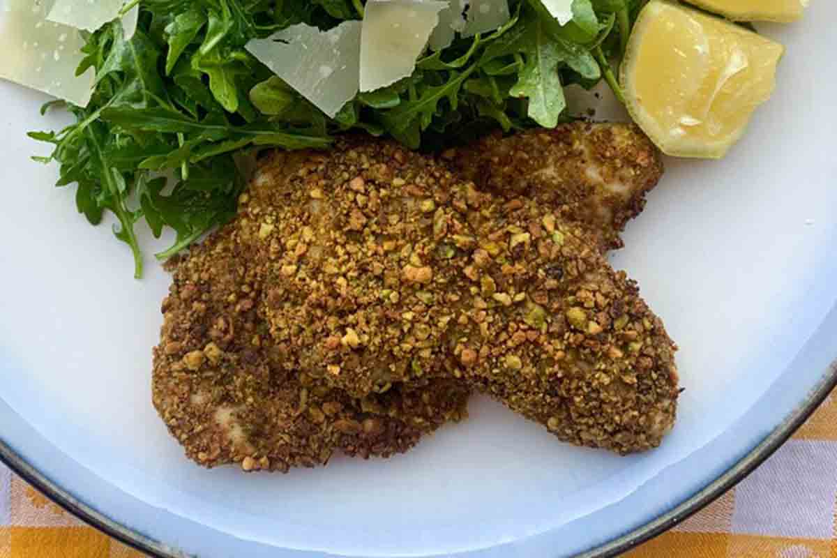 Two pistachio-crusted chicken cutlets on a white plate with an arugula and Parmesan salad on the side.
