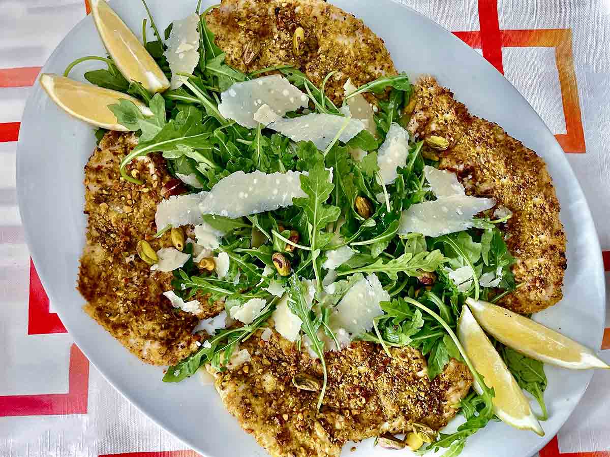 Four pistachio-crusted chicken cutlets on a white platter, topped with arugula, Parmesan cheese, lemon wedges, and pistachios.
