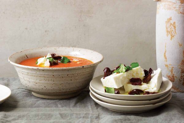 A ceramic bowl filled with chilled tomato soup with feta and olives and a stack of three white plates topped with feta, olives, and basil.