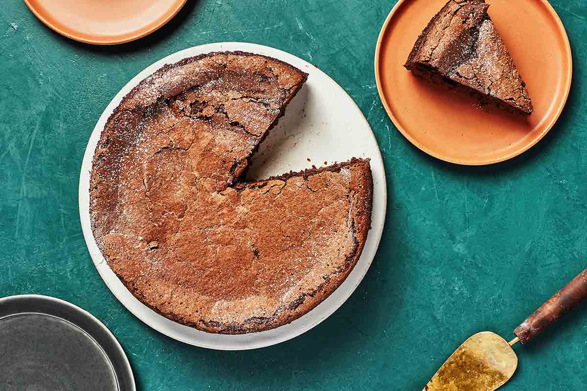 A round chocolate polenta pudding cake on a white plate with one slice cut from it on an orange plate nearby.