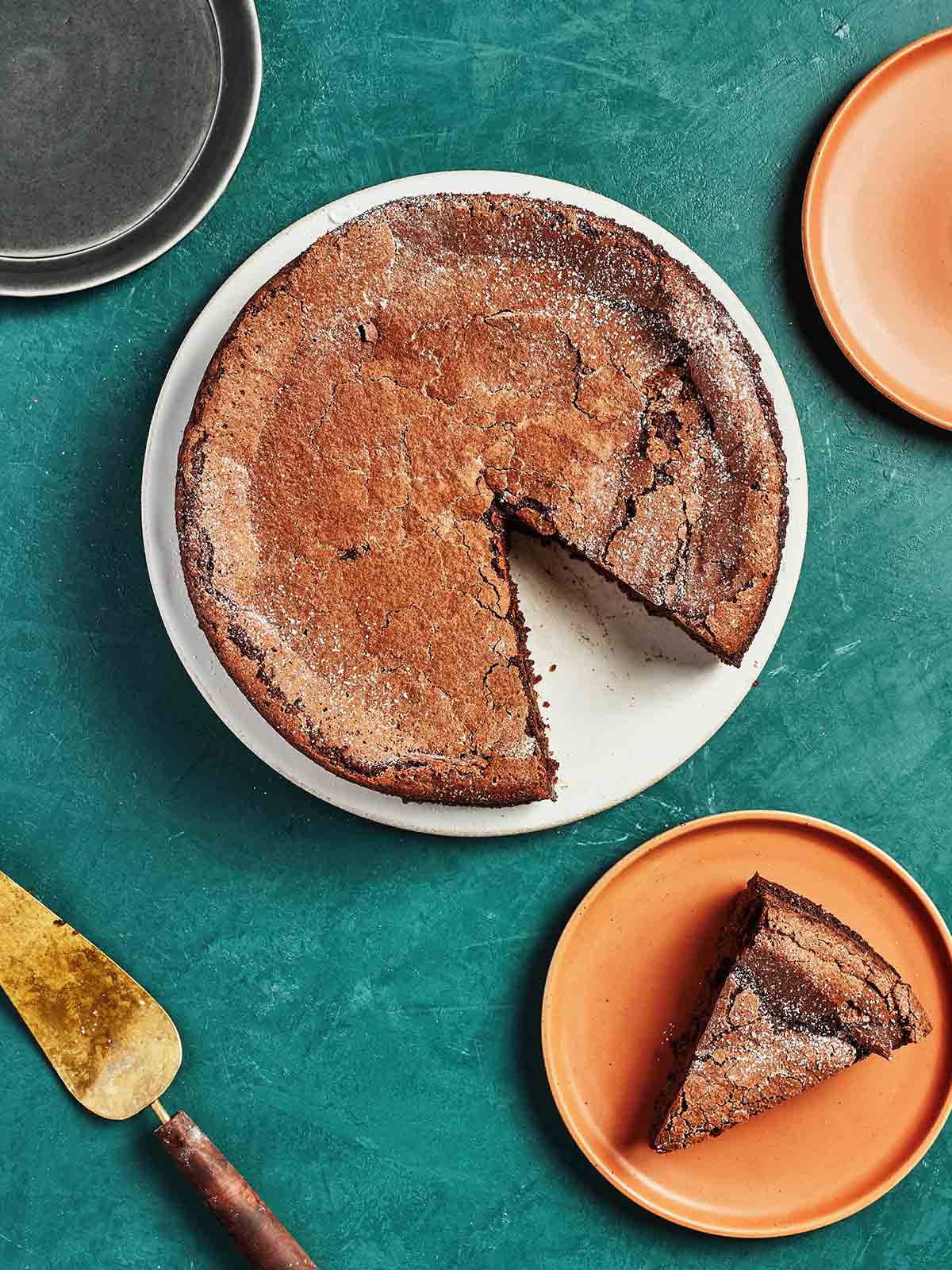 A round chocolate polenta pudding cake on a white plate with one slice cut from it on an orange plate nearby.