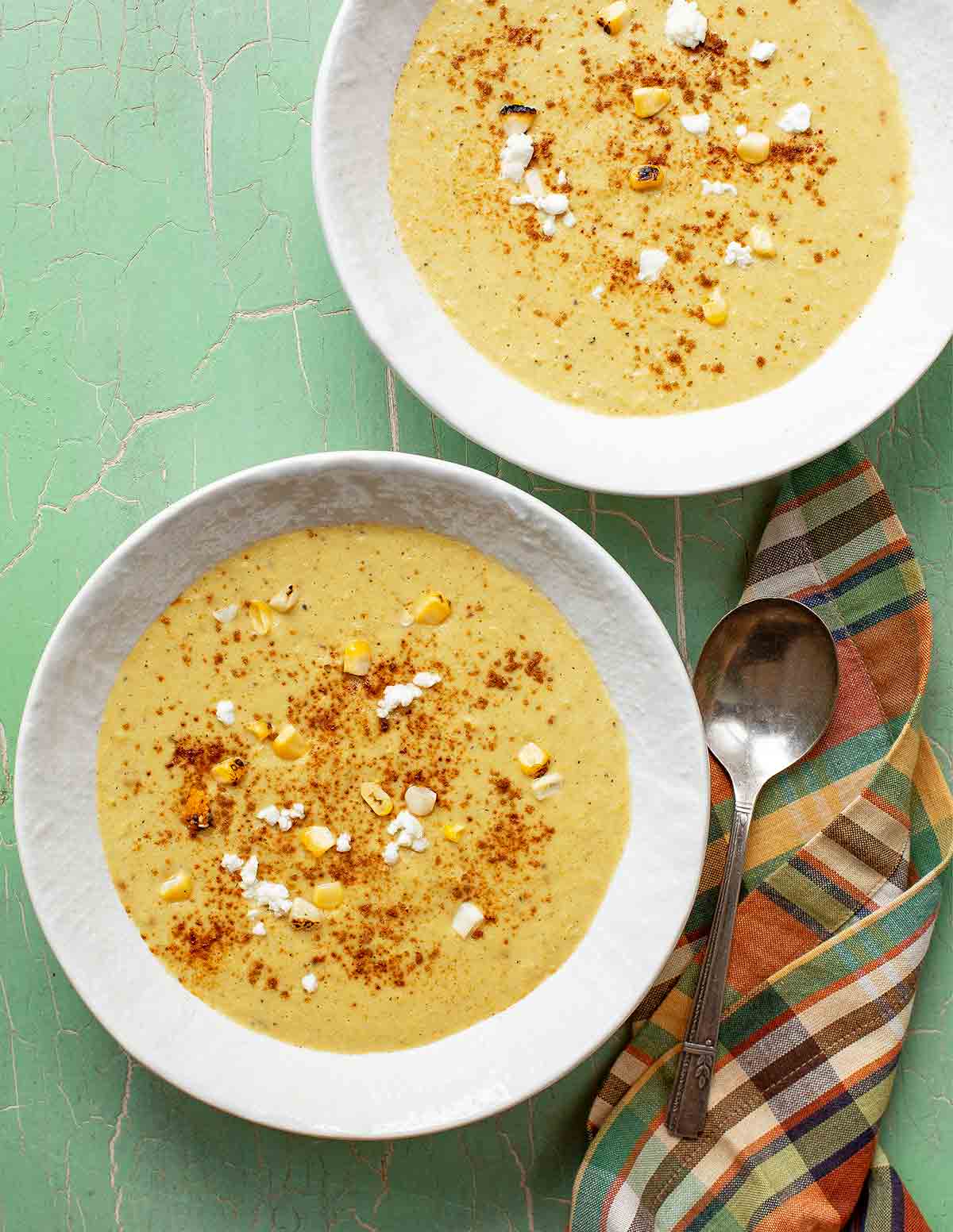 Two white bowls of corn and roasted poblano soup garnished with cheese, paprika, and corn kernels, with a napkin and spoon on the side.