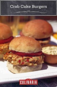 A platter with several crab cake burgers topped with tomato in a soft roll.