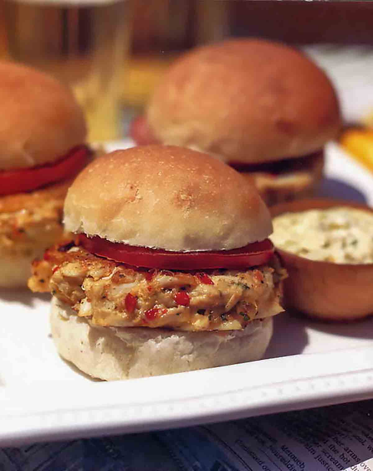 A platter with several crab cake burgers topped with tomato in a soft roll.