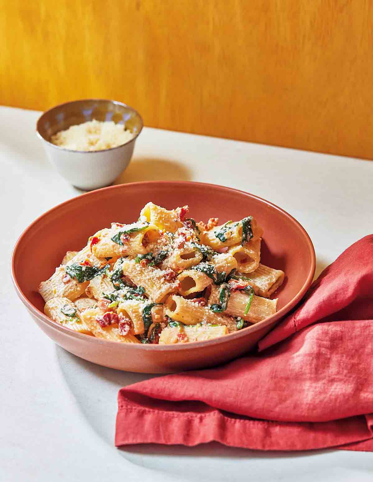 A red bowl filled with creamy sun-dried tomato and spinach pasta with a red cloth napkin on the side.