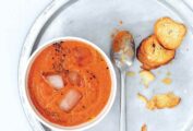 A bowl of easy gazpacho with ice cubes floating in it on a silver plate with a spoon and a few crostini.