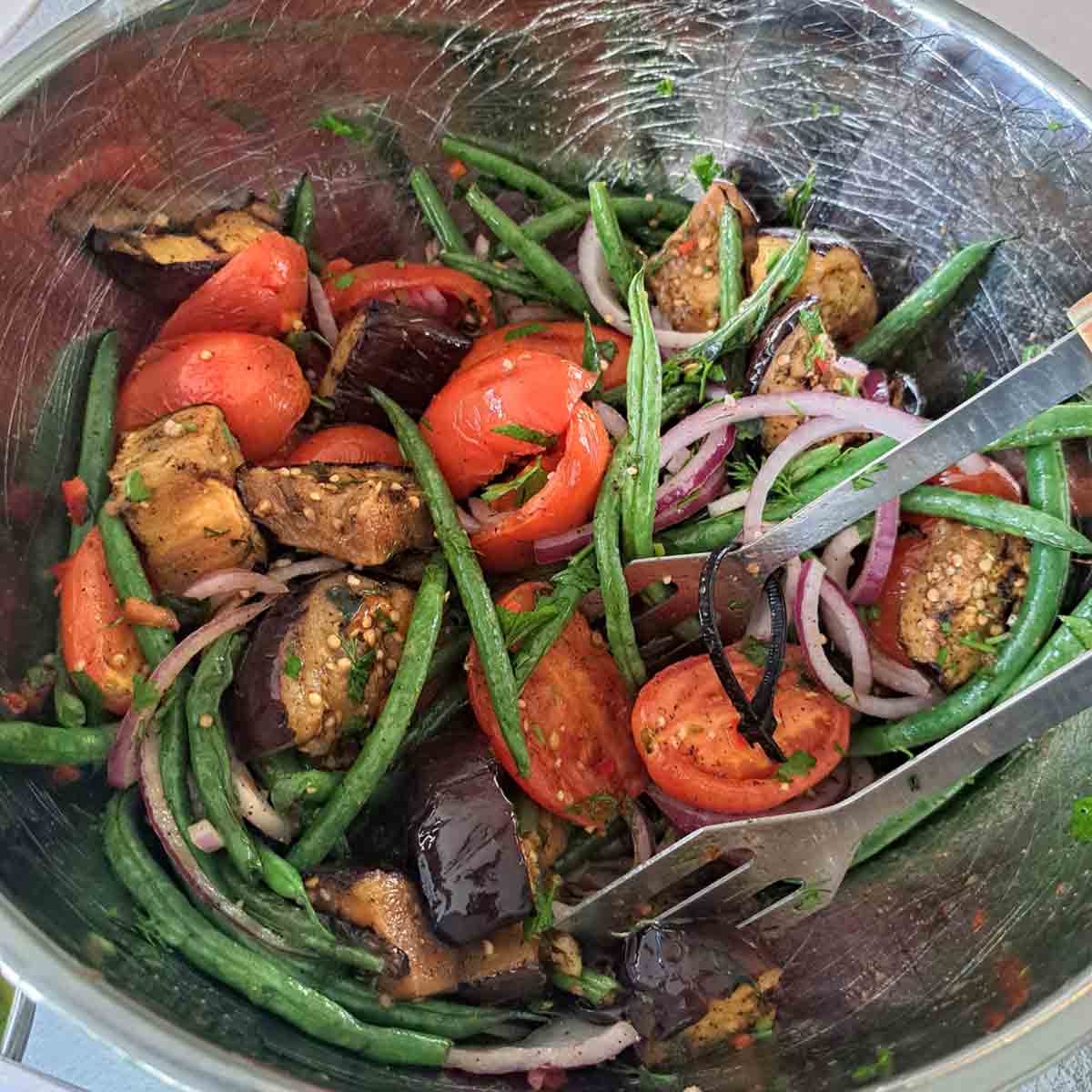 Grilled tomatoes, beans, eggplant, and onions in a metal bowl with serving tongs.