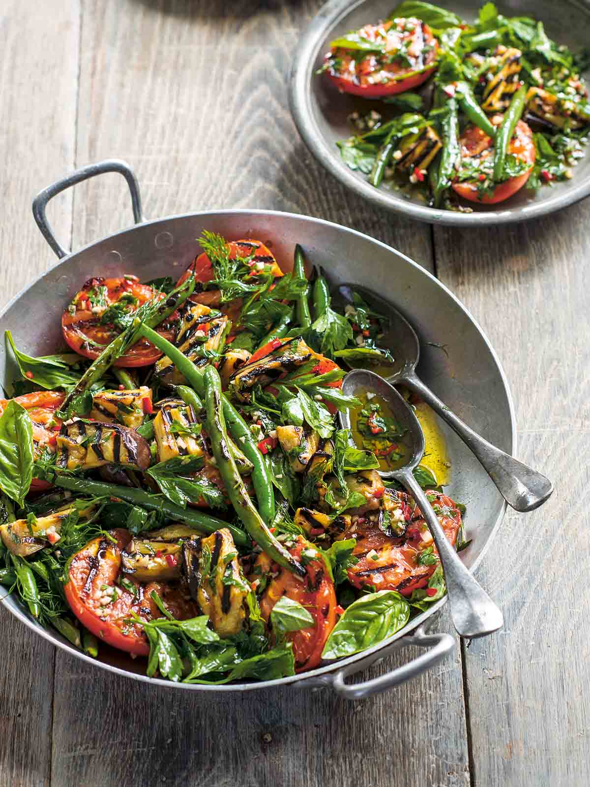 A large and small bowl filled with grilled vegetables salad and two serving spoons in the large bowl.