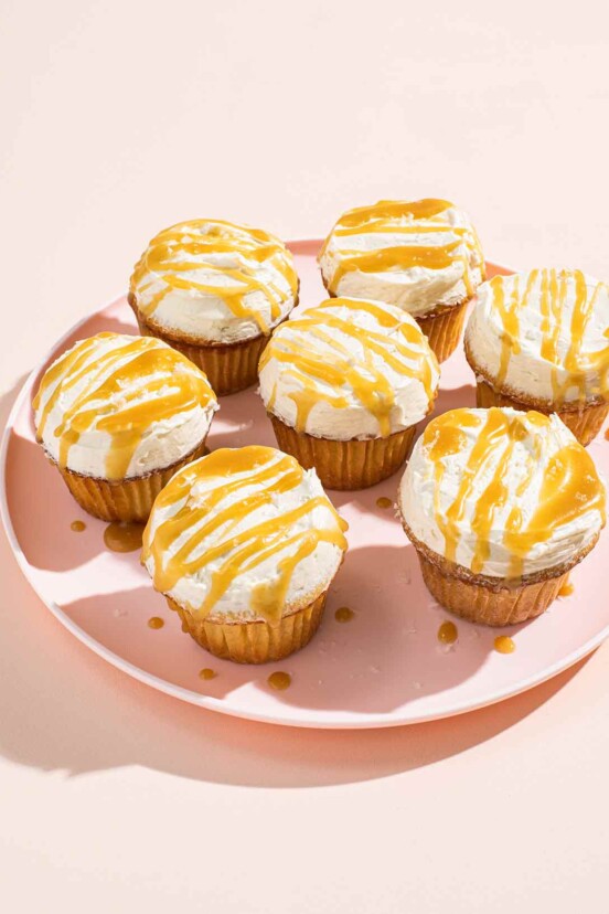 Seven frosted cupcakes with butterscotch drizzled on top on a pink plate.