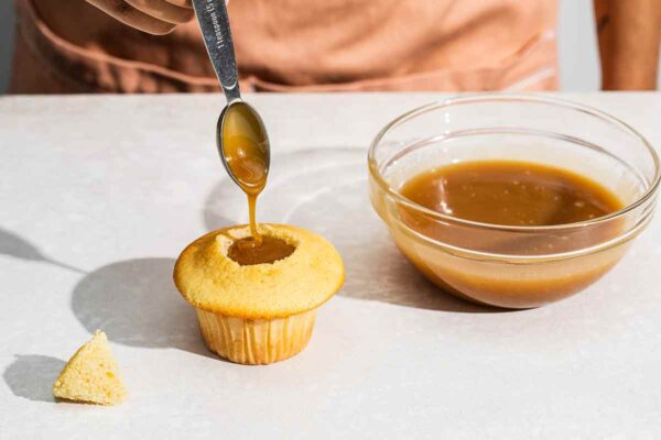 A person scooping butterscotch sauce from a glass bowl into the center of a cupcake.