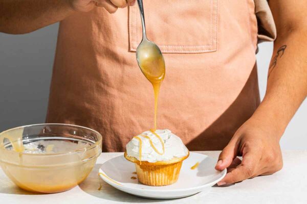 A person drizzling butterscotch sauce from a bowl over a frosted cupcake.
