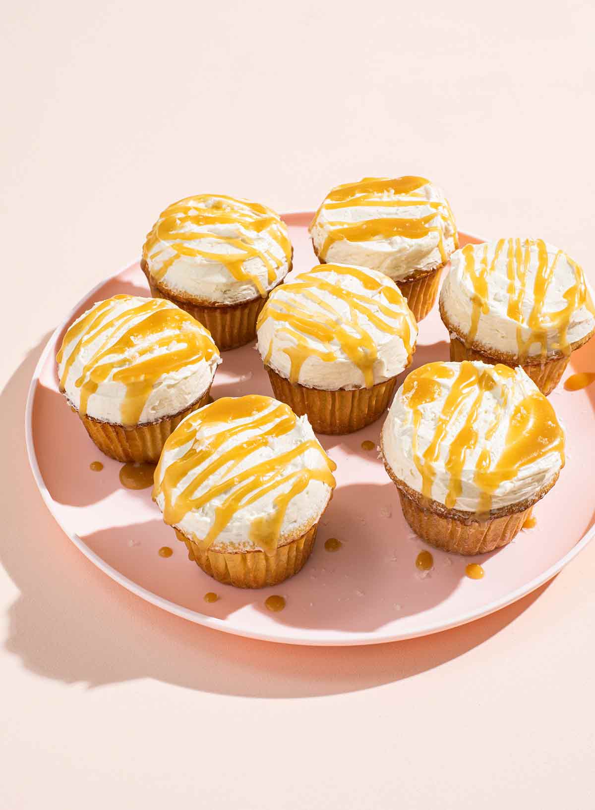 Seven frosted cupcakes with butterscotch drizzled on top on a pink plate.
