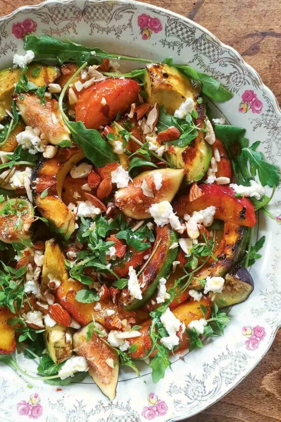 A decorative platter filled with grilled avocado, grilled peach slices, fresh figs, herbs, and goat cheese.