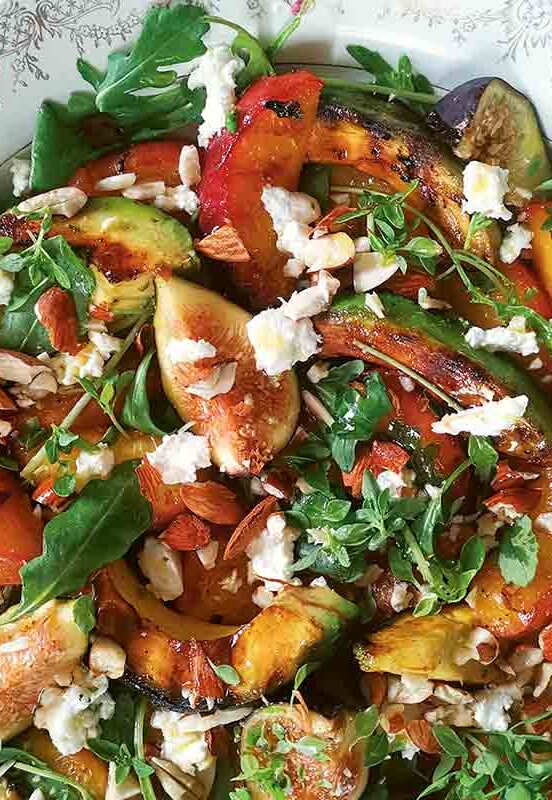 A decorative platter filled with grilled avocado, grilled peach slices, fresh figs, herbs, and goat cheese.
