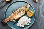 A whole grilled fish on a plate with a scoop of creamy cucumber salad and half of a grilled lemon.
