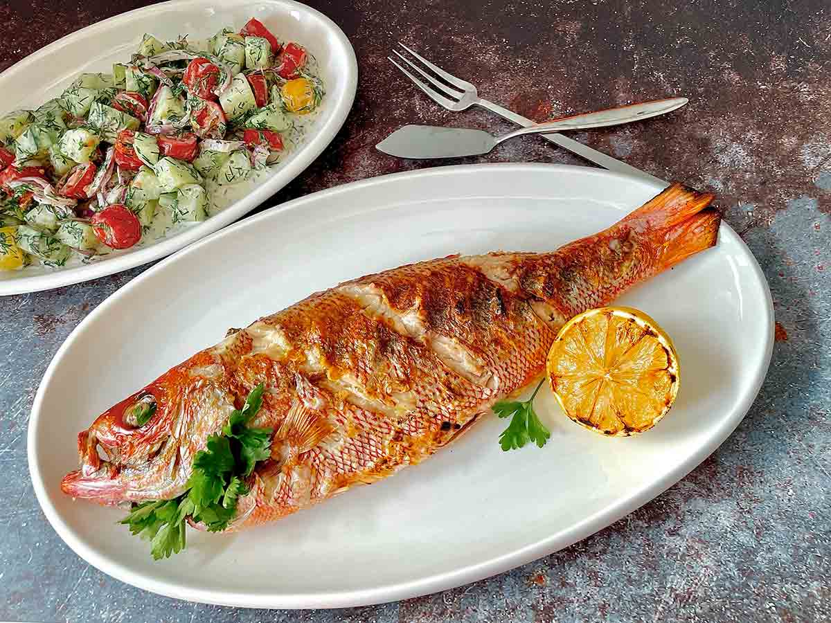 A grilled whole fish on a white platter with a grilled lemon half and a creamy cucumber salad on a separate platter.