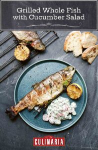 A whole grilled fish on a plate with a scoop of creamy cucumber salad and half of a grilled lemon.