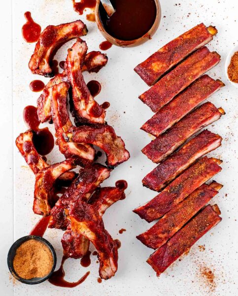Two racks of ribs cut into individual ribs, one spice-rubbed and one sauced, with bowls of spice rub and bbq sauce on the side.