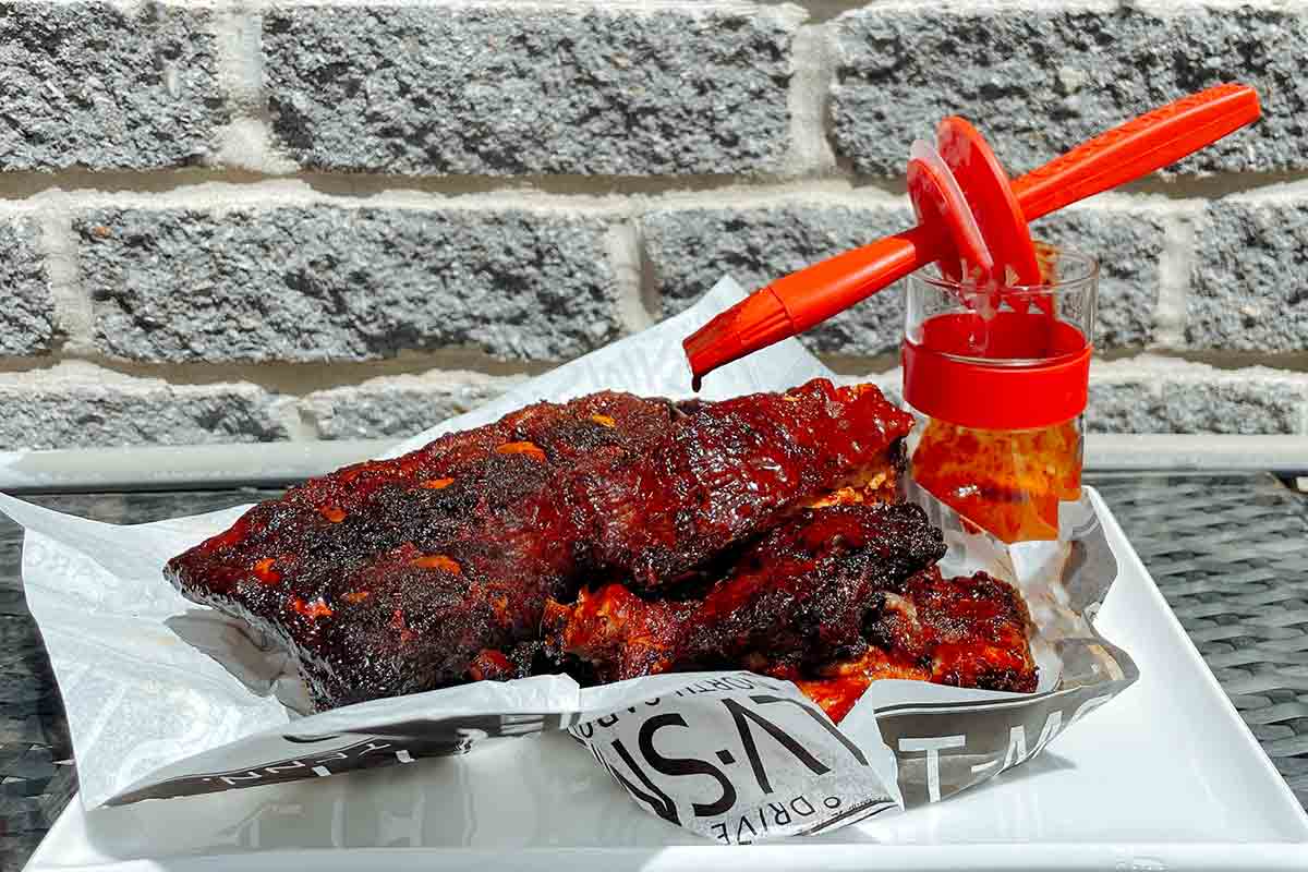 Two racks of smoked pork ribs on a piece of serving parchment on a white plate with a basting brush and sauce jar resting above.