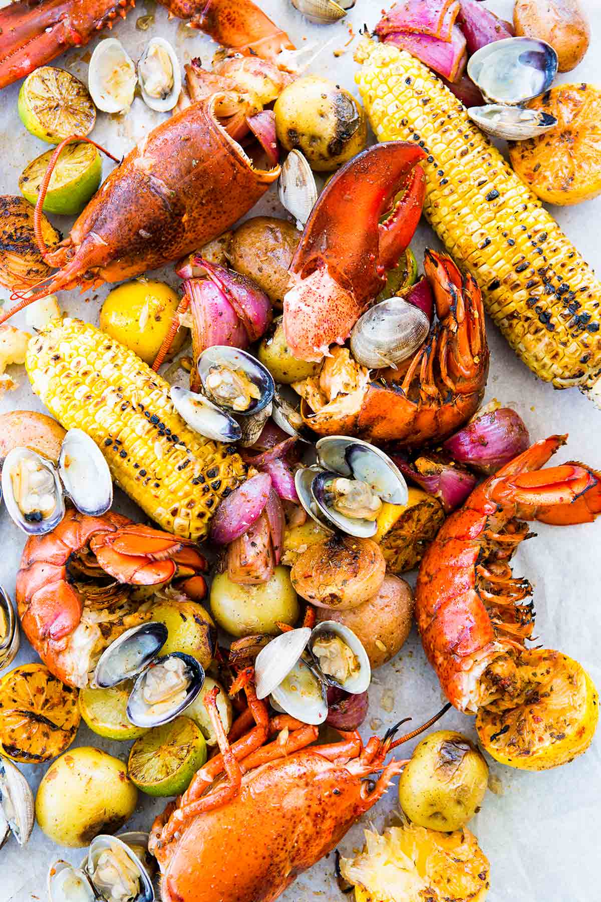 All the fixings for a New England clambake - lobster, clams, potatoes, corn, lemon - scattered on a white surface.