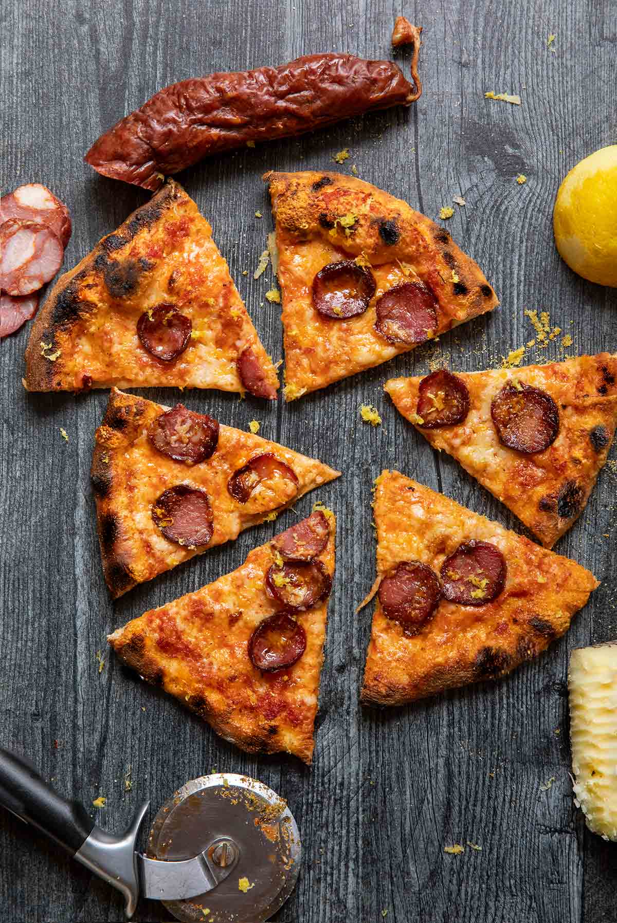 An Ooni pizza with Portuguese sausage and cheese on gray weathered wood, with chouriço, lemon, cheese, and pizza cutter.