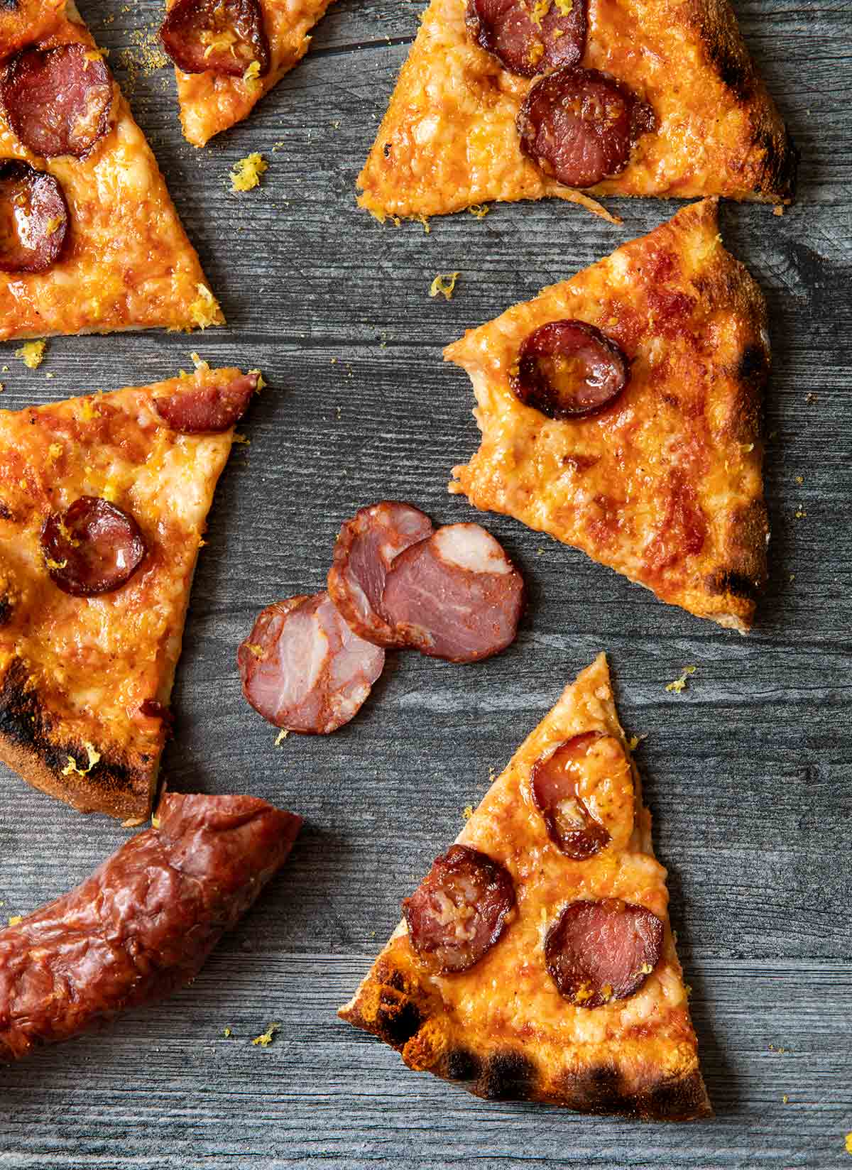 Sic slices An Ooni pizza with Portuguese sausage and cheese on gray weathered wood, with chouriço.
