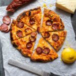 An Ooni pizza with Portuguese sausage and cheese on gray weathered wood, with chouriço, lemon, cheese, and pizza cutter.