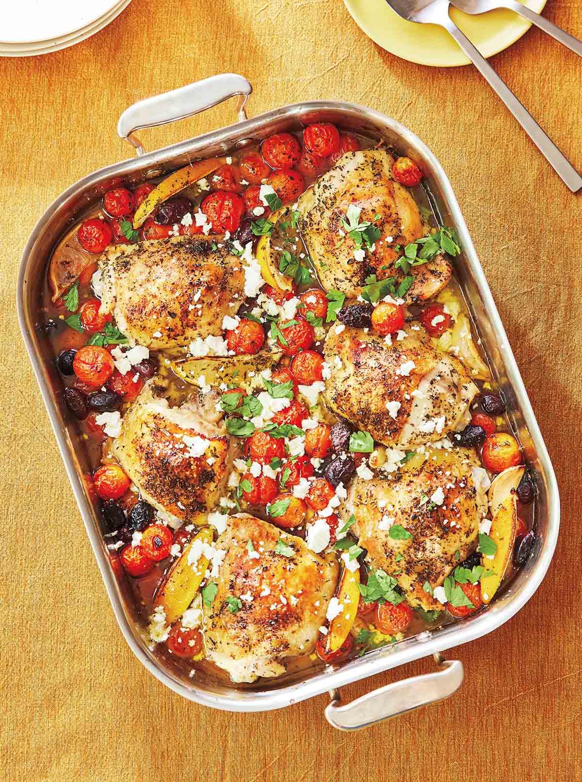 A metal roasting pan filled with roasted chicken thighs, olives, lemon, tomatoes, feta, and herbs.