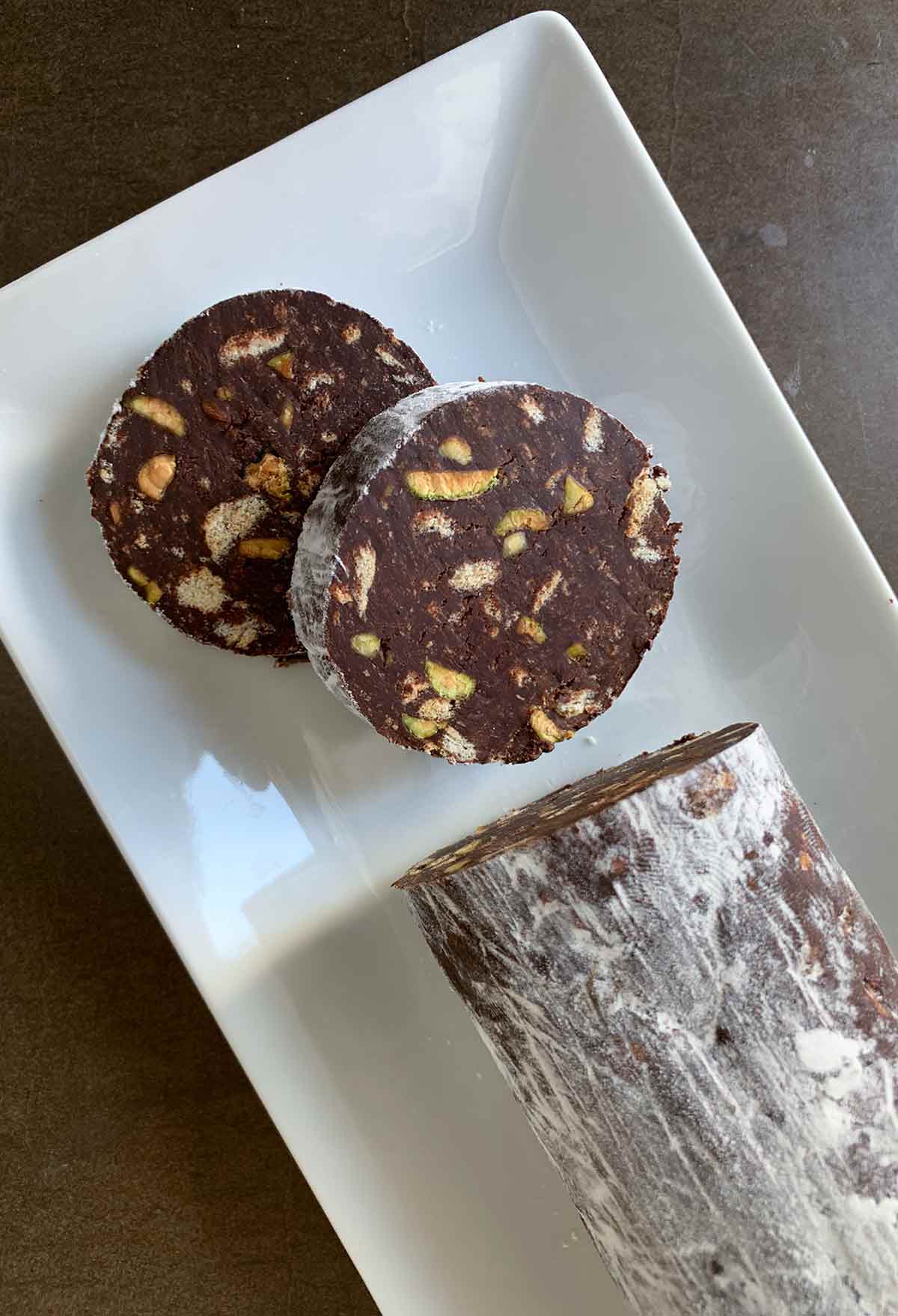 A log of chocolate salami with two slices cut from the end on a white rectangular platter.